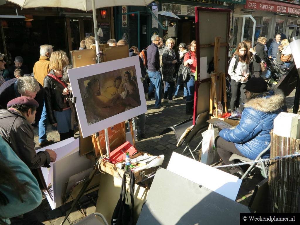 De toeristen lopen langs de rand van de markt. Bij deze kraam kun je schilderijen kopen. De schilderijen worden overigens speciaal voor toeristen gemaakt, en soms in China.